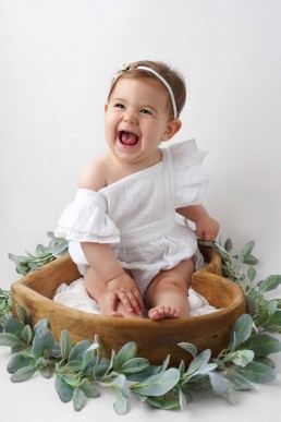 A baby girl in a white romper sits in a wooden bowl.