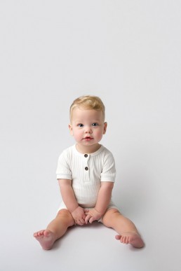 A baby in a white romper sitting on a white background.