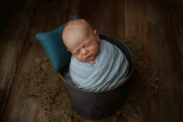 A newborn peacefully sleeping in a bucket on a wooden table for newborn photography in Essex.
