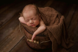 A newborn peacefully sleeping in a bucket on a wooden floor for an Essex photoshoot.
