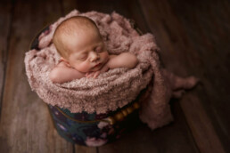 Capture stunning images of a newborn peacefully sleeping in a bucket on a wooden floor. Perfect for newborn photography in Essex.