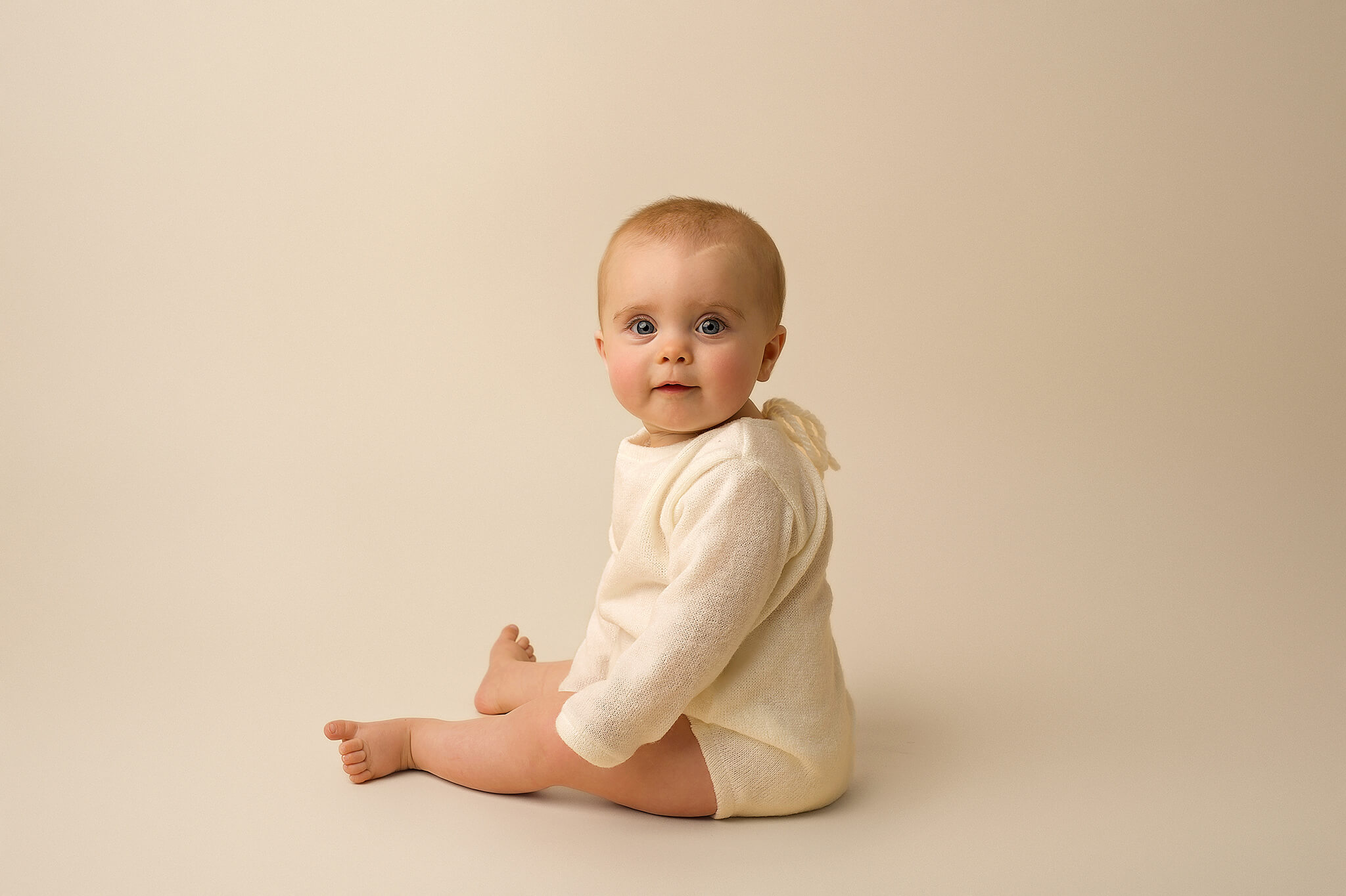 A baby sitting on the floor in a white outfit.