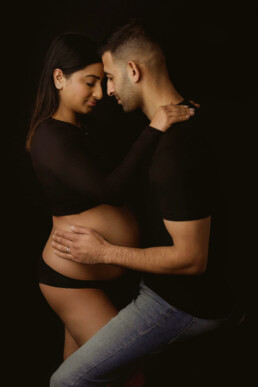 A pregnant couple posing in front of a black background.