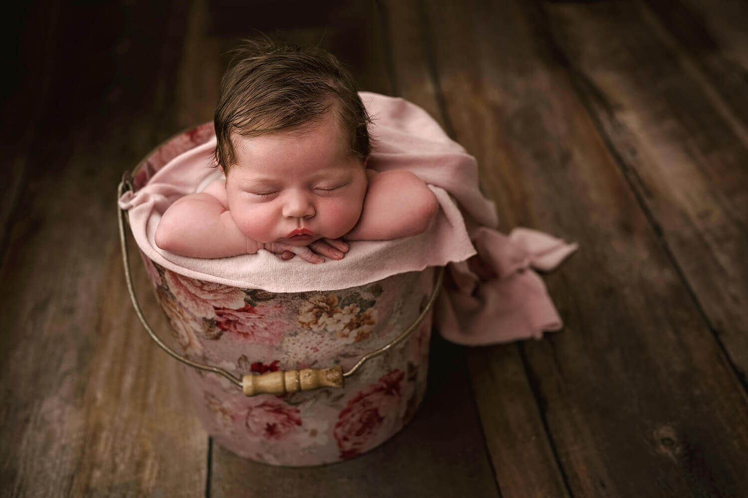 Capture precious moments with professional newborn photography in Essex. Watch as your little one peacefully sleeps in a bucket on a wooden floor, creating timeless memories to cherish forever.