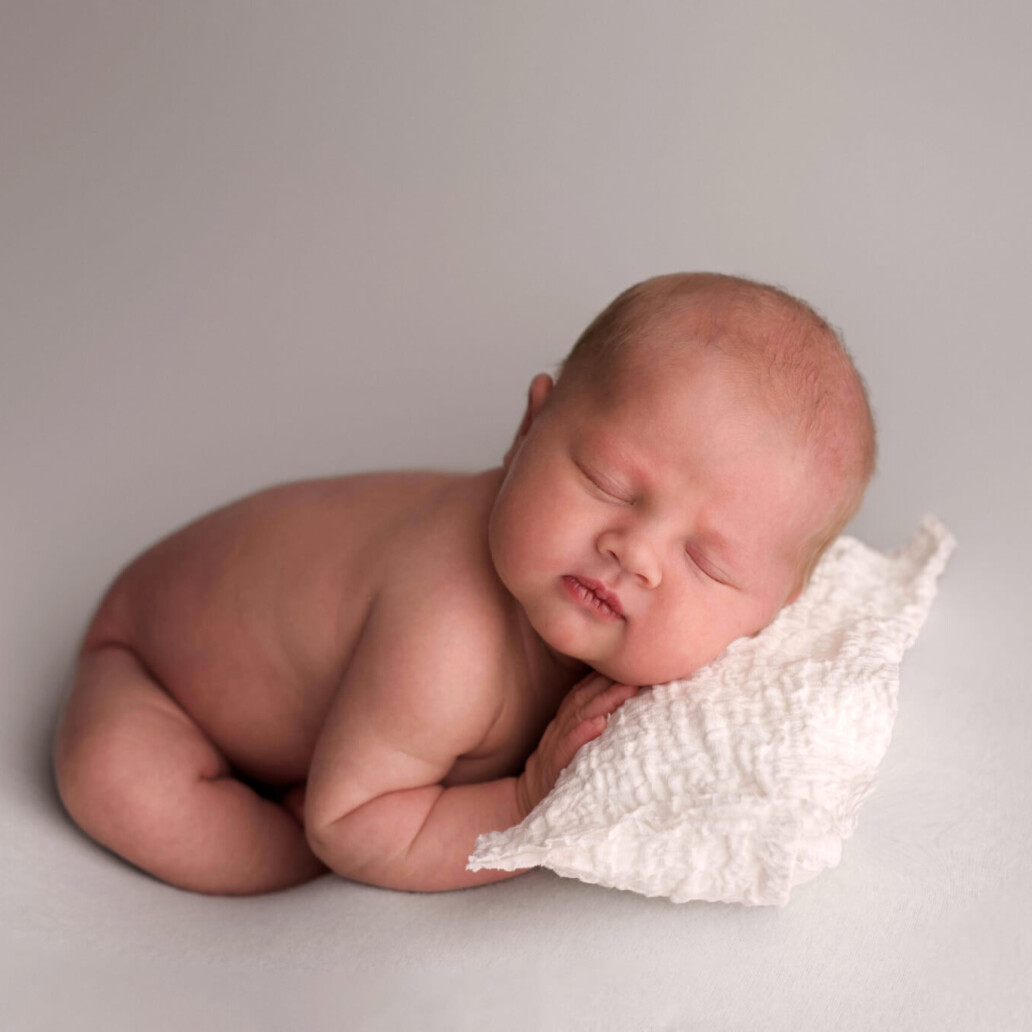 A sleeping baby rests on a soft, textured white pillow against a plain background.