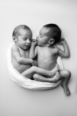 Two newborn babies lie closely together, wrapped in a soft white blanket. They are facing each other with eyes closed, peacefully sleeping.