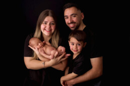 A family of four poses against a black background. The parents hold a newborn, and a young child stands beside them, all wearing black clothing.