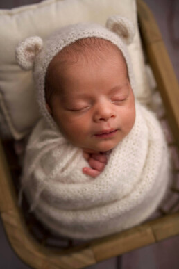 Newborn baby peacefully asleep, wrapped in a creamy soft blanket, wearing a matching knit hat with bear ears, resting in a cushioned basket.