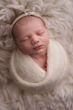 Newborn baby wrapped in a cream blanket, sleeping peacefully on a fluffy white surface.