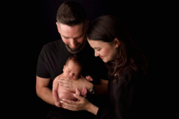 A man and woman gently cradle a newborn baby, all wearing black clothing, against a black background.