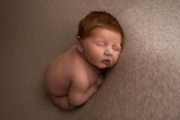 A newborn baby with closed eyes is posed asleep on a soft, neutral-toned blanket, facing sideways.