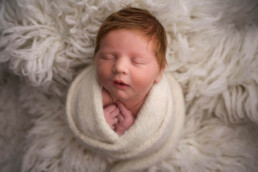 Newborn baby wrapped in a cream-colored blanket, sleeping peacefully on a soft, textured white surface.