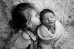 A young girl kisses a swaddled newborn baby on a soft, fluffy surface. The scene is in black and white.