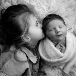 A young girl kisses a swaddled newborn baby on a soft, fluffy surface. The scene is in black and white.