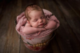 A sleeping baby is tucked in a floral bucket lined with a pink blanket, positioned on a wooden floor.