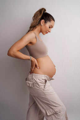 Pregnant woman in a beige top and light pants stands sideways, hand on hip, looking down at her belly against a plain background.