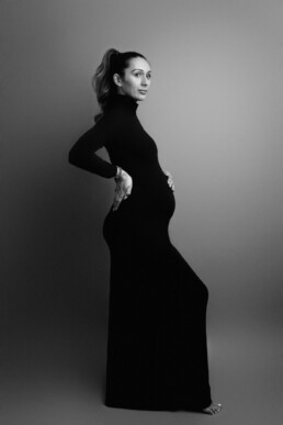 A person in a form-fitting black dress poses in profile against a plain background, showcasing a visible baby bump.