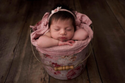 A sleeping baby with dark hair rests in a floral bucket, wrapped in a pink blanket, on a wooden floor.