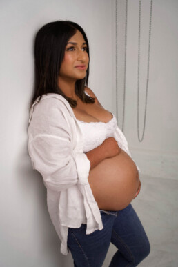 A pregnant woman in a white shirt and jeans leans against a white wall, holding her bare belly and looking slightly upwards.