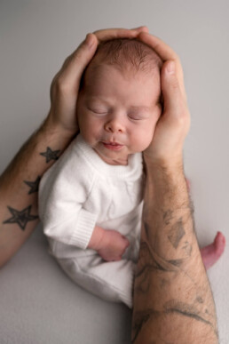 A newborn baby in a white outfit is cradled gently by tattooed adult hands; the baby has closed eyes and a peaceful expression—capturing the beauty of newborn photography Essex.