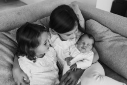 Three young children, including a newborn, sit closely together on a couch. The older children hold the infant gently. Capturing this tender moment in black and white, the image beautifully exemplifies the timeless style of newborn photography Essex families adore.