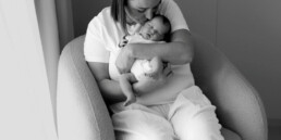 Woman sitting in an armchair, holding and cuddling a sleeping baby in her arms near a window in a softly lit room—a serene moment beautifully captured, perfect for newborn photography Essex.