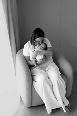 Woman sitting in an armchair, holding and cuddling a sleeping baby in her arms near a window in a softly lit room—a serene moment beautifully captured, perfect for newborn photography Essex.