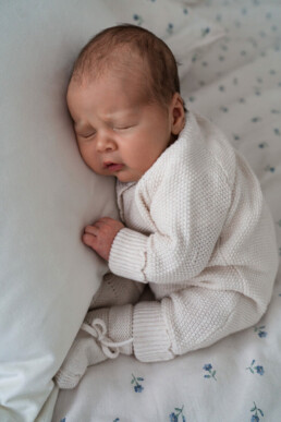 Newborn baby in a cream knit outfit sleeps on a white pillow and patterned sheets, lying on their side with eyes closed.
