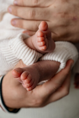 Close-up of an adult’s hands gently holding a baby’s feet, with the baby wearing a white knit outfit—capturing a tender moment in newborn photography Essex.