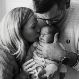 A woman kisses a sleeping baby held by a man who gently cradles the baby&rsquo;s head. All three are close together, creating an intimate family moment.