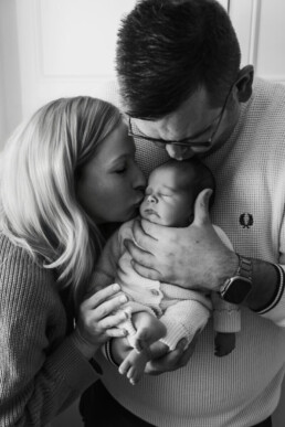 A woman kisses a sleeping baby held by a man who gently cradles the baby’s head. All three are close together, creating an intimate family moment.
