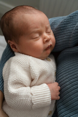 A newborn baby wearing a white knitted sweater is sleeping, being held by someone in a blue knitted garment.