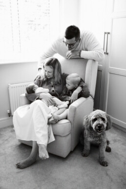A woman sits on a chair holding a baby and a toddler, while a man stands behind her and a dog sits on the floor nearby, capturing an intimate family moment typical of newborn photography Essex style.