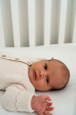 A baby wearing a cream knit sweater lies on a white sheet in a crib, looking up with one arm outstretched.
