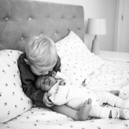 Toddler sits on a bed and kisses the forehead of a baby lying beside them. Both children are on patterned bedding in a bedroom.