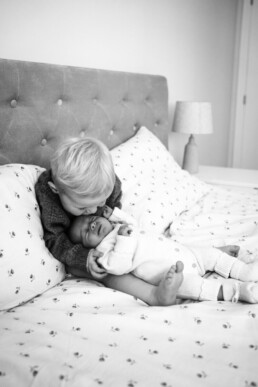 Toddler sits on a bed and kisses the forehead of a baby lying beside them. Both children are on patterned bedding in a bedroom.