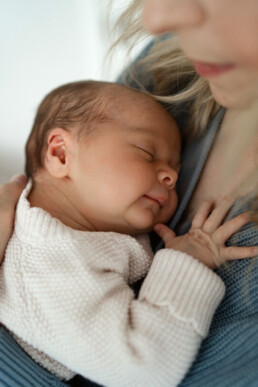 A newborn baby sleeps peacefully in the arms of an adult wearing a blue sweater. The adult’s face is partially visible, and the baby’s hand rests on the adult’s chest.