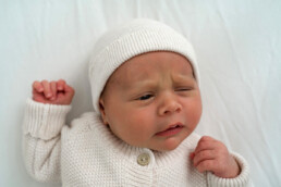 A newborn baby in a white knit outfit and hat lies on a white surface, with one eye open and a slightly furrowed brow&mdash;capturing the gentle innocence often sought after in newborn photography Essex.