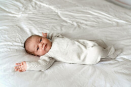 Newborn baby in a light-colored onesie lies on a white bed with arms outstretched, looking up&mdash;an adorable moment perfectly captured for newborn photography Essex.