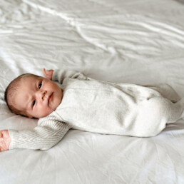 Newborn baby in a light-colored onesie lies on a white bed with arms outstretched, looking up&mdash;an adorable moment perfectly captured for newborn photography Essex.